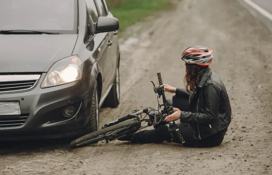 A cyclist in a helmet sits on the road beside her bike, in front of a car