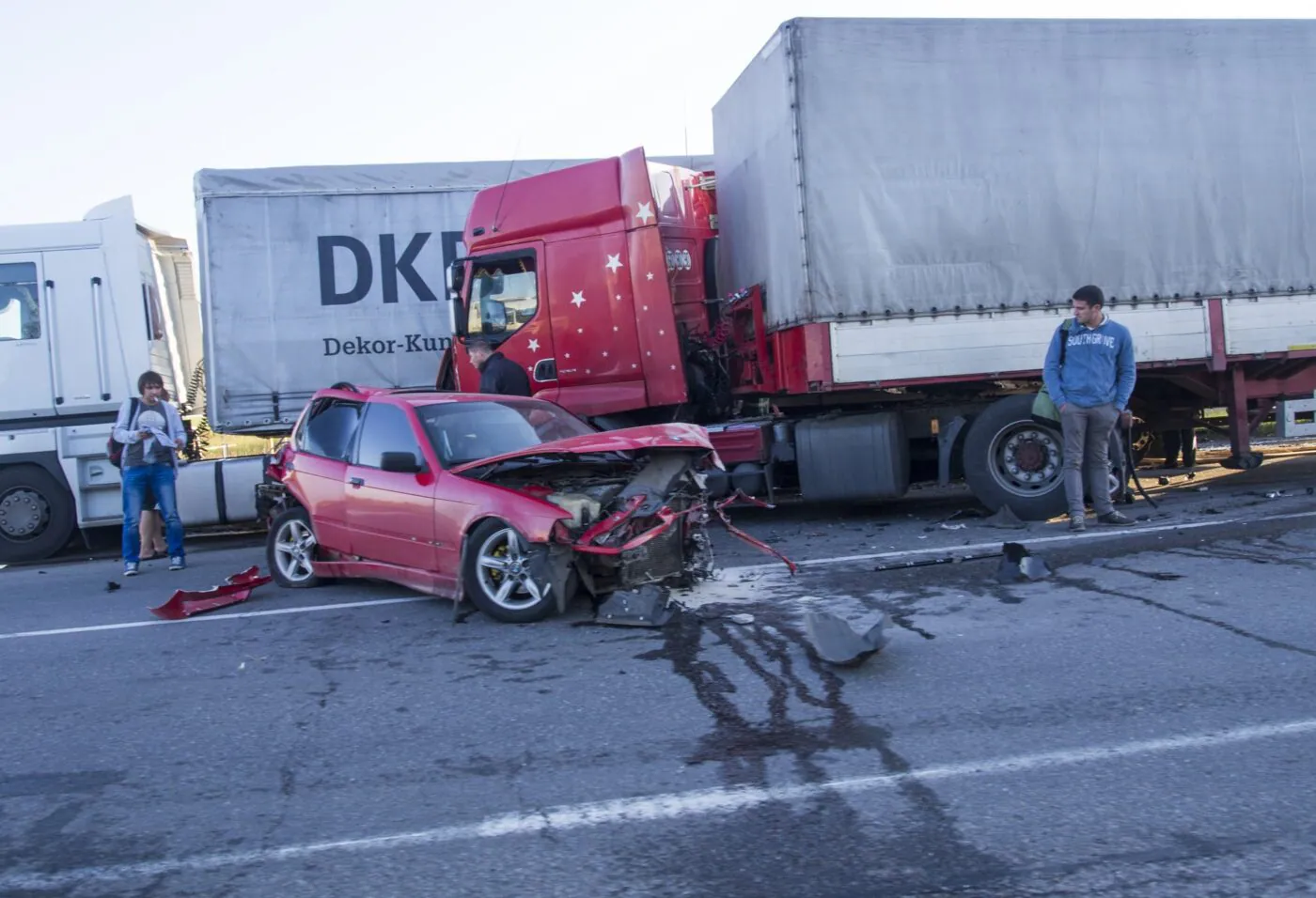 A red car crushed against the back of a tractor-trailer as two people look on