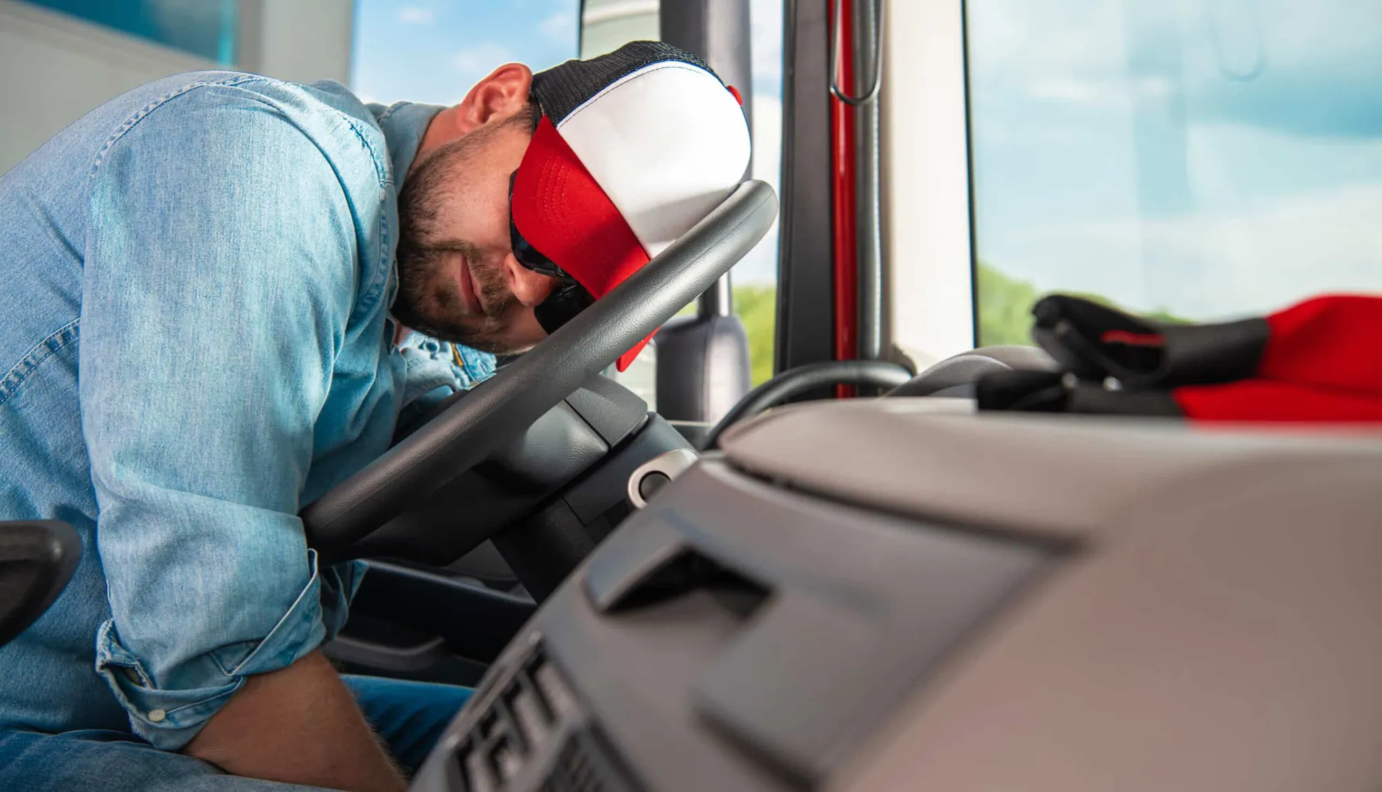 A truck driver in a cap and sunglasses slumped asleep over the steering wheel in the cab