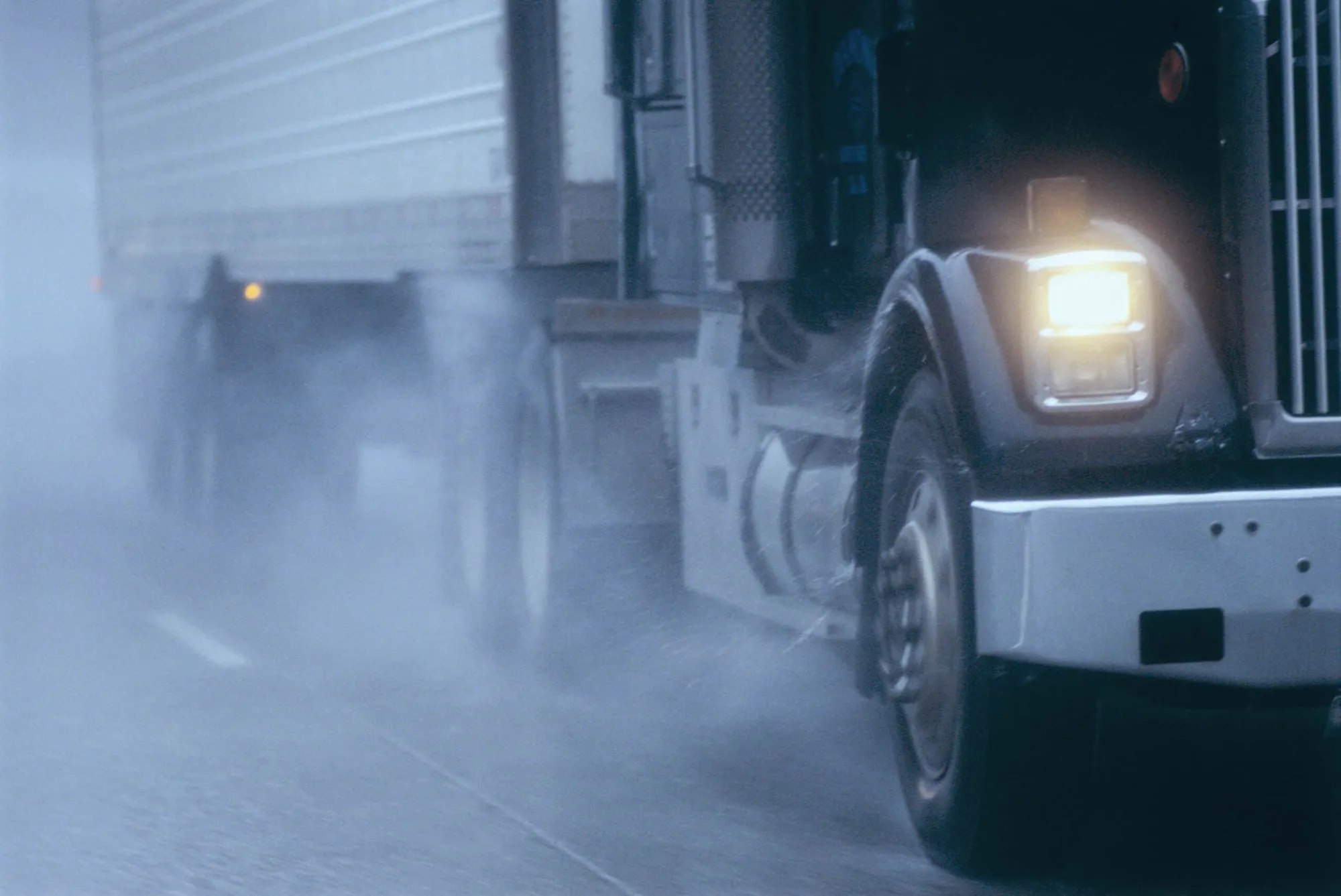 A tractor-trailer driving through rain and road spray with its headlights on