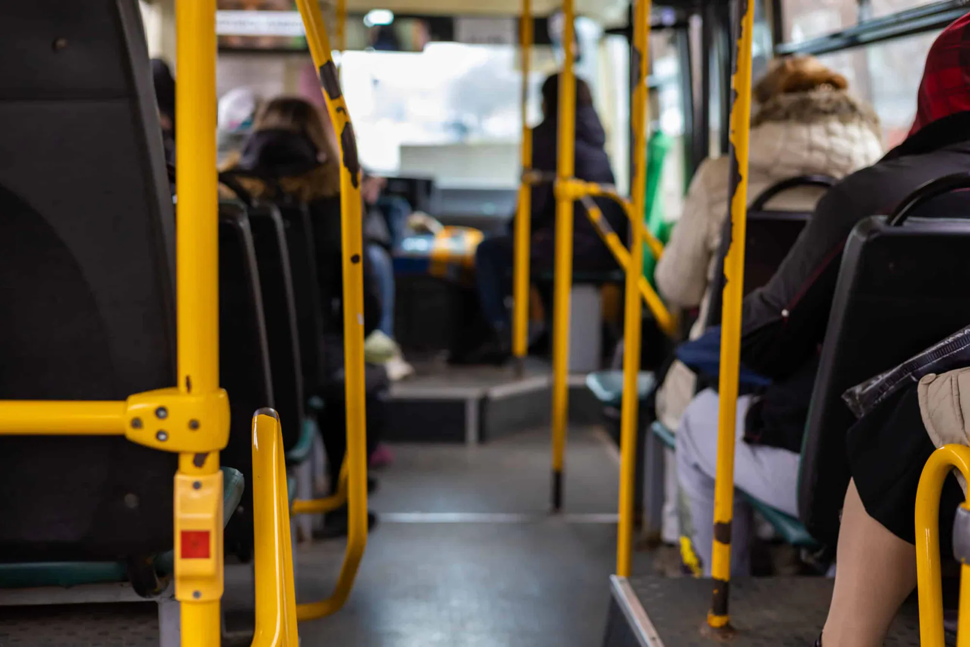 Passengers seated on a city bus, seen from the aisle between yellow handrails