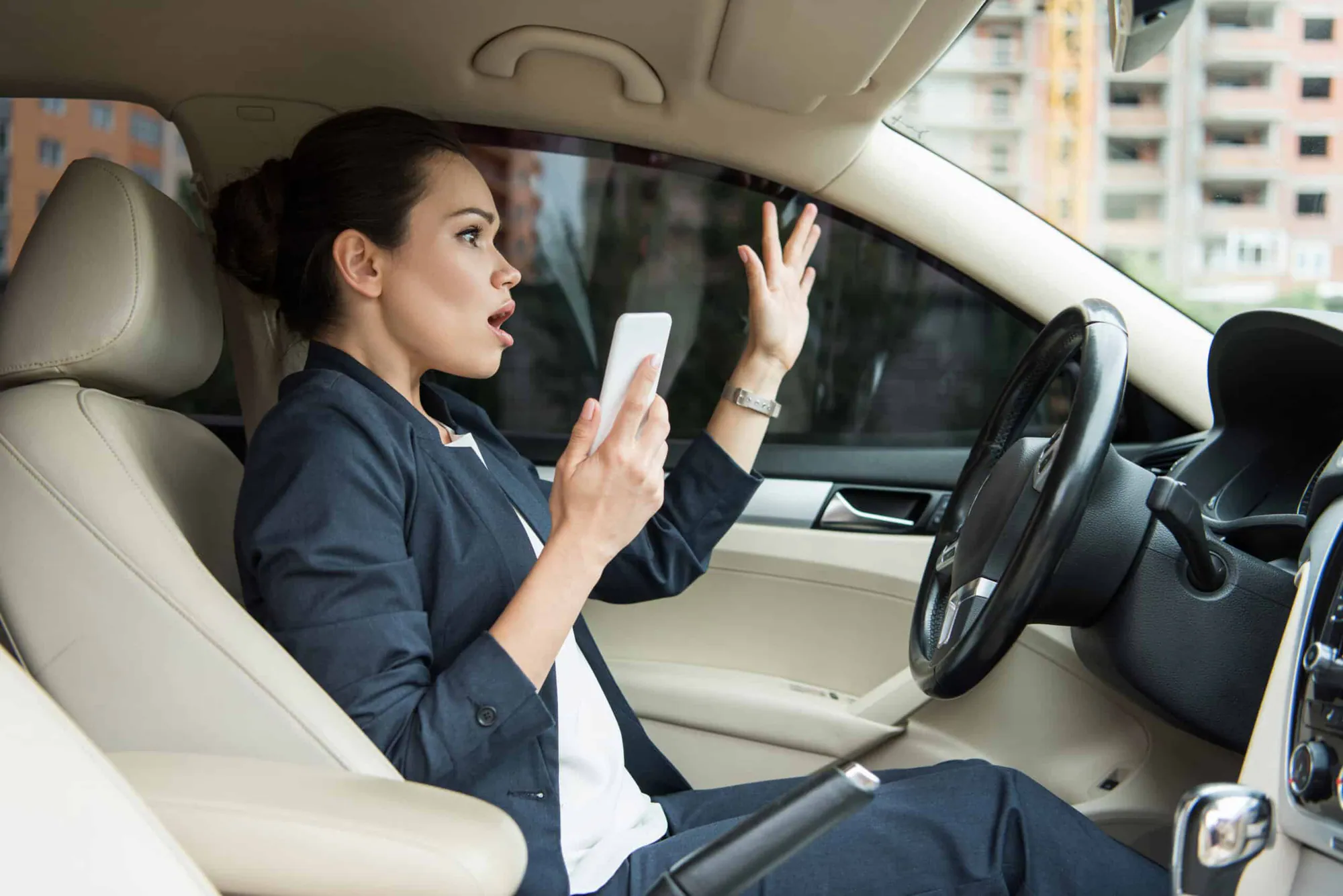 A driver holds a phone in one hand and raises the other in alarm behind the wheel