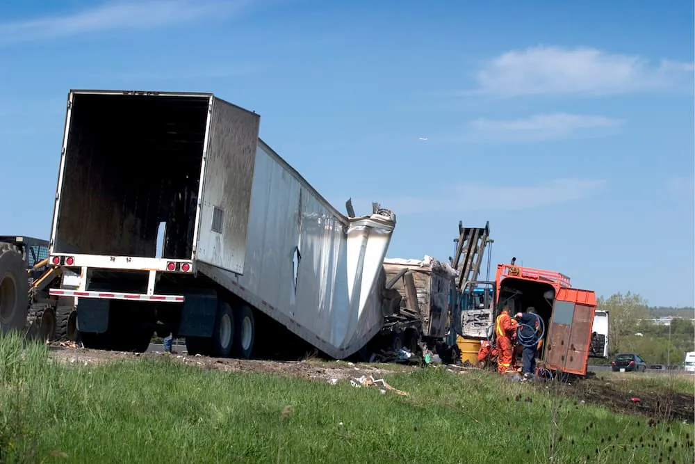 A wrecked tractor-trailer with its trailer torn open beside a highway while a recovery crew works