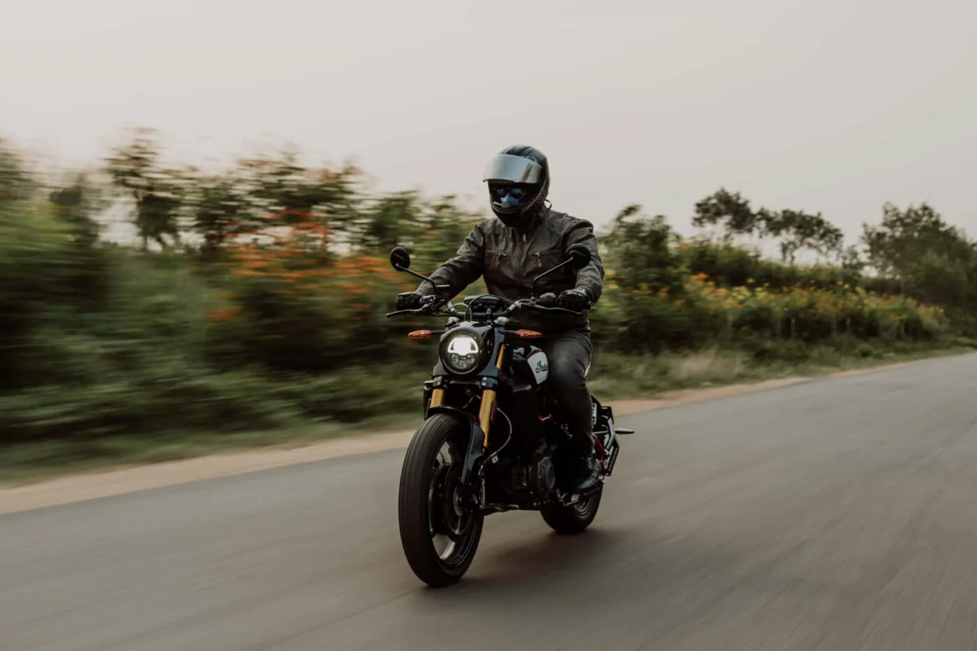 A rider in a full-face helmet and leather jacket rides a motorcycle down a two-lane road