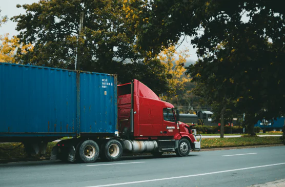 A red semi truck hauling a blue shipping container along a tree-lined road