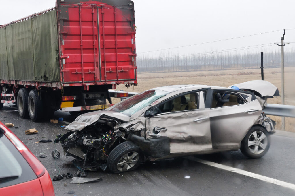 Silver sedan with a crushed front end after colliding with a red 18-wheeler on a wet highway